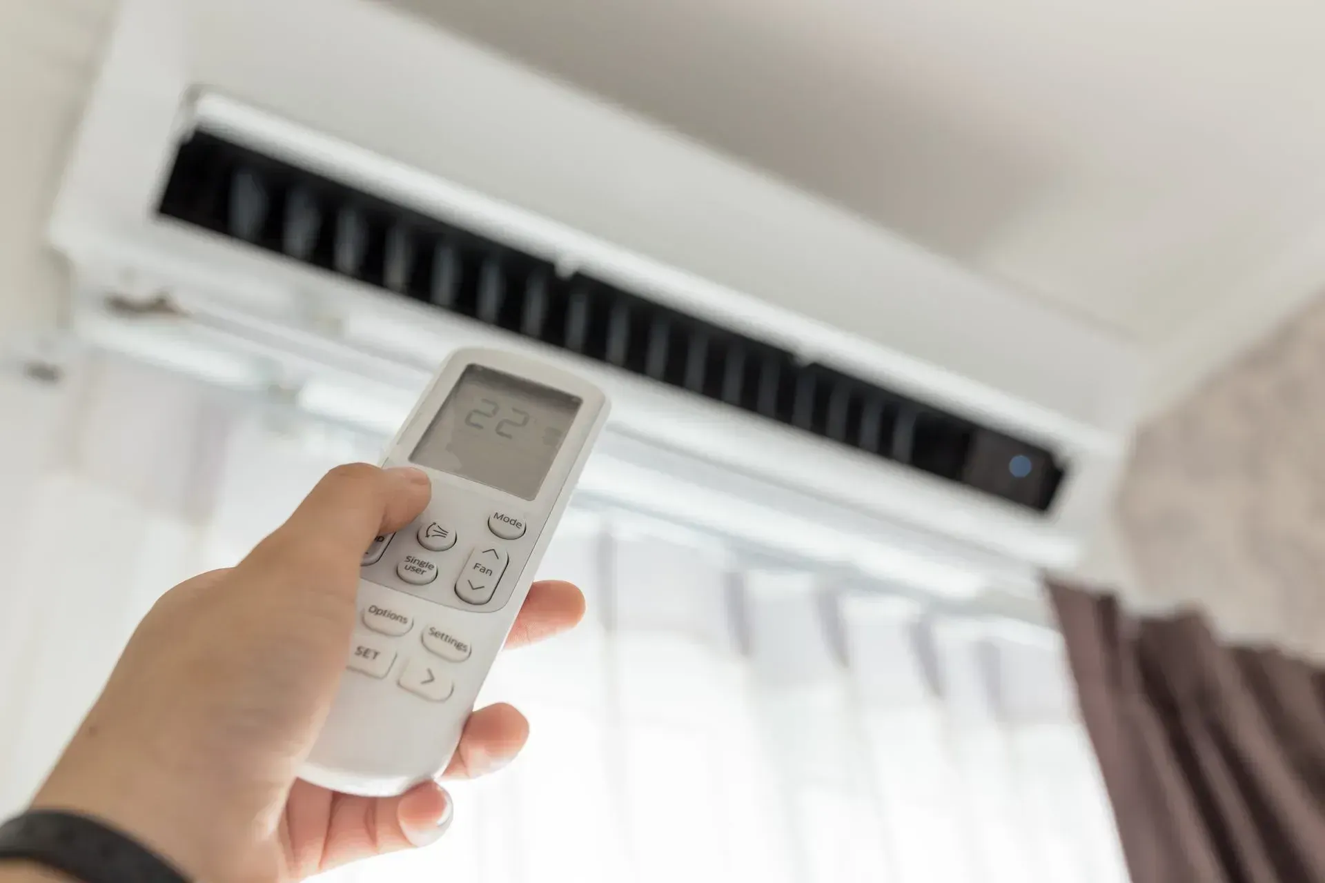 Hand holding a remote control in front of a wall-mounted air conditioner.