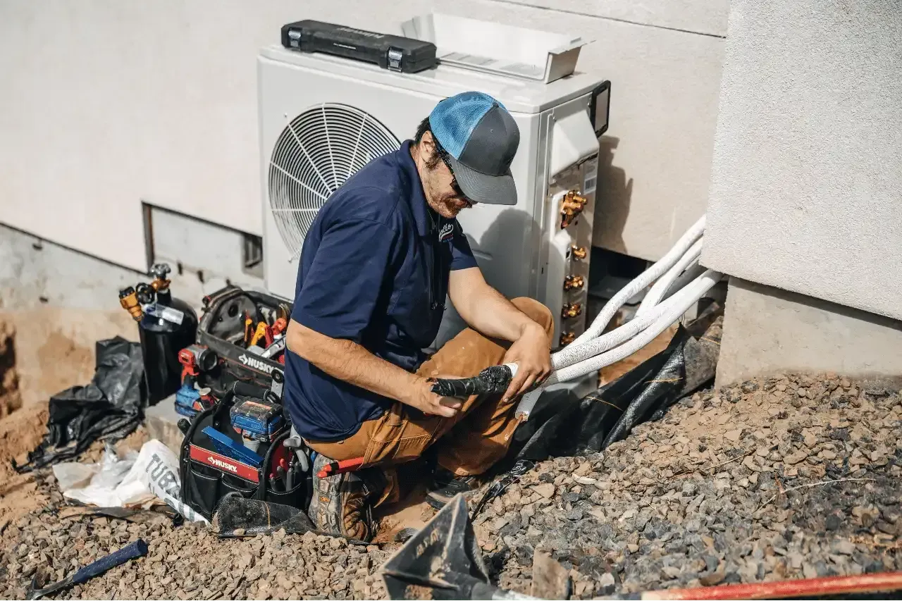 HVAC technician kneeling beside an outdoor condenser unit with tools laid out.